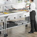 A man and woman using a ServIt electric steam table with a sneeze guard in a school kitchen.