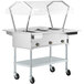 A ServIt stainless steel electric steam table with a clear sneeze guard over hot food on wheels.