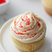 A cupcake with white frosting and Regal Red nonpareils on a white surface.