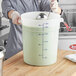 A woman uses a measuring cup to pour liquid into a large white Vigor food storage container on a counter.