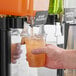 A hand holding a plastic cup of orange liquid in front of a green Pure Craft Beverages beverage dispenser.