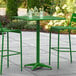A Lancaster Table & Seating green bar table with two green chairs on an outdoor patio.