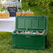 A Hunter green CaterGator outdoor cooler filled with ice and bottles of soda on a grassy area.