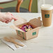 A hand reaching for a salad in a New Roots Kraft paper take-out box with a wooden spoon and fork on a white napkin.