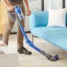 A person using a blue and silver Lavex Pro cordless stick vacuum to clean a couch.