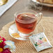 A glass cup of Bigelow Rose and Mint herbal tea with a tea bag in it on a saucer with a packet of tea.