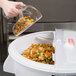 A hand pouring multicolored spiral pasta from a clear container into a white Rubbermaid ingredient bin.