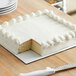 A white square cake on a white corrugated board with a slice missing.