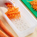 A person pouring small pieces of orange carrots from a wooden surface into a translucent plastic container over a Vigor 1/3 size drain tray.