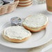 A white plate with a bagel and a portion cup of Philadelphia Chive and Onion Cream Cheese Spread.