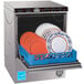 A CMA undercounter dishwasher with a rack of white plates with blue and orange designs.