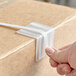 A person's hand holding a white Lavex poly strap attached to a cardboard box using a metal edge protector.