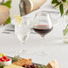 A Della Luce Maia burgundy wine glass filled with red wine on a table next to a plate of food.
