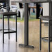 A black Lancaster Table & Seating bar table base on a table with a cup and a chair.