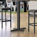 A black Lancaster Table & Seating bar height table base with a table and two chairs.