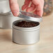 A hand opening a silver metal tin filled with brown tea leaves.