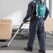 A man in a blue uniform using a Makita backpack vacuum to clean a room.