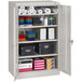 A Tennsco light gray storage cabinet with solid doors filled with boxes and rolls of paper.