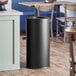 A black Lancaster Table & Seating decorative waste receptacle on a wood floor.