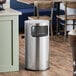 A Lancaster Table & Seating stainless steel round decorative trash can on a wood floor.