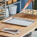 A metal baking sheet pan sits on a wooden countertop with a pair of tongs on a cloth nearby, while a person holds another pan with unbaked pastries in the background.