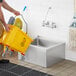 A man using a yellow bucket to fill a stainless steel mop sink.