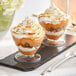 Two Choice clear plastic dessert cups with whipped cream and caramel on a white tablecloth.