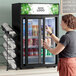 A woman opening an Avantco black countertop display refrigerator with a sliding glass door to take a beverage.