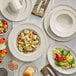 An Acopa Condesa warm gray porcelain pasta bowl with a scalloped edge on a table with plates and a bowl of fruit salad.