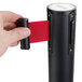 A hand holding a red retractable belt attached to a black Aarco crowd control stanchion.