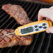 A hand holding a yellow Taylor digital pocket probe thermometer to a piece of meat on a grill.