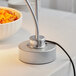 A silver Avantco weighted base for a bulb warmer heat lamp over food on a hotel buffet table.