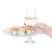 A hand holding a CAC Bright White china plate with a sushi roll on it and a glass of liquid.