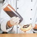 A person using a Tablecraft Dualway squeeze bottle to pour chocolate sauce onto a doughnut.