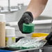 A person wearing black gloves holding a green enzymatic grease trap block near cleaning supplies.