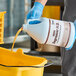 A person in blue gloves pouring Noble Eco Degreaser liquid into a yellow bucket.