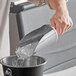 A hand using a Choice stainless steel flour scoop to pour ice into a metal container.