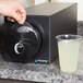 A hand using a San Jamar black countertop lid dispenser to put a lid on a glass of lemonade.