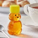 A small plastic bear shaped bottle of honey with a yellow cap next to a cup of tea.