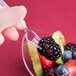 A hand holding a WNA Comet clear plastic tasting fork over a bowl of blackberries.