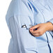 A woman wearing a sky blue Uncommon Chef long sleeve chef coat with a mesh back and putting a cell phone in the pocket.