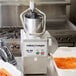 A Robot Coupe food processor with a full moon pusher processing carrots.