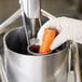 A gloved hand places a carrot in a Robot Coupe food processor.