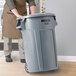 A person in an apron pushing a Rubbermaid BRUTE 44 gallon grey trash can.