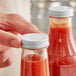 A hand holding a bottle of red sauce with a white metal lid.