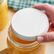 A hand holding a 89/400 white metal lid over a jar of yellow liquid.