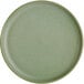 An Acopa Pangea sage matte coupe porcelain plate with a speckled texture on a green surface.