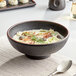 A close up of an Acopa Heika black matte stoneware bowl filled with seafood and greens.