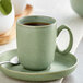A sage green Acopa Pangea porcelain cup and saucer with a spoon on a saucer.