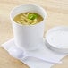 A white Huhtamaki paper food cup filled with soup and noodles with a white vented lid and a spoon.